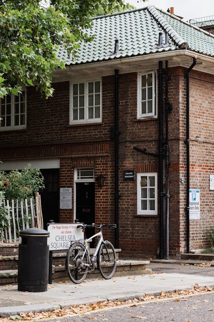 A two-story brick building with a pitched, grey tiled roof and white-framed sash windows on both floors is visible in the image. The building's façade features dark guttering and downpipes, with some black pipework visible against the red brick wall. In front of the building, on the pavement, there is a black cylindrical trash bin positioned near the curb. A white bicycle with a black saddle and handlebars is parked upright, leaning against a small white signboard that displays the words 'Chelsea Square' and provides some additional information, partially obscured. The surrounding environment includes a leafy green tree extending its branches partly over the scene, casting some shade on the building and pavement. To the right of the bicycle, a blue and white parking restriction sign indicates designated parking zones, and the pavement is scattered with fallen leaves, suggesting an autumn setting. The overall scene appears to relate to a residential or mixed-use area where private waste collection may be arranged outside designated authority collection times, emphasizing the importance of independent rubbish removal services like Rubbish Removal Chelsea for maintaining cleanliness and managing waste efficiently.