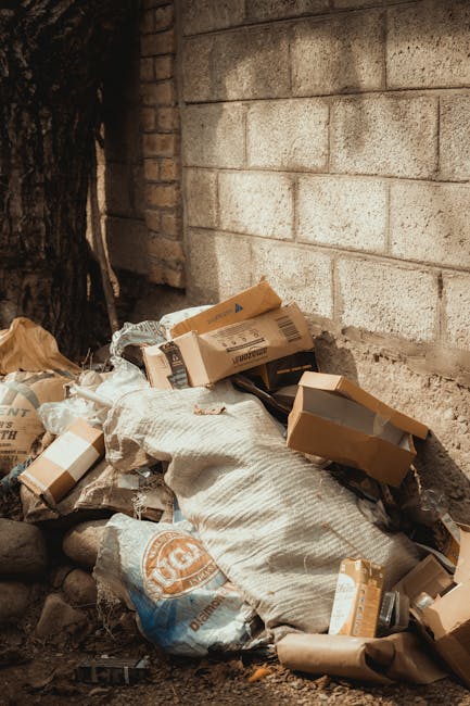 A pile of miscellaneous rubbish, including cardboard boxes, plastic bags, and paper packaging, is stacked against a concrete brick wall outdoors. The cardboard boxes vary in size and shape, with some flattened while others remain intact, showing printed labels and logos. The plastic bags appear crumpled and are partially filled, containing unknown materials. A large woven sack is also visible, contributing to the clutter. The rubble is positioned on uneven ground with small stones and dirt surrounding it. To the left, a tree trunk with dark, textured bark is partially visible, casting slight shadows over the rubbish. The background features a weathered concrete wall with visible texture and slight discoloration, suggesting an exterior setting. Overhead lighting creates soft shadows, emphasizing the textures and shapes of the discarded waste. This scene aligns with the type of private or alternative rubbish collection services offered by Rubbish Removal Chelsea, particularly in the context of on-site clearance or non-municipal disposal of waste.