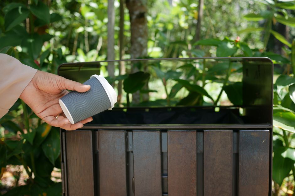 A person's hand, wearing a beige long-sleeve garment, is holding a grey disposable coffee cup with a white lid, positioned above an open, black metal trash bin. The bin's interior is empty, with rusted and weathered metal sides. The scene is set outdoors, surrounded by lush green foliage, including tall plants with broad leaves, in natural daylight that creates soft shadows and highlights the textures of the leaves and metal surfaces. The background features dense greenery with sunlight filtering through the foliage, providing a natural and vibrant environment typical of outdoor waste disposal areas, where independent rubbish collection and disposal practices might occur, as exemplified by the presence of a personal coffee cup and a publicly accessible trash bin for waste removal.