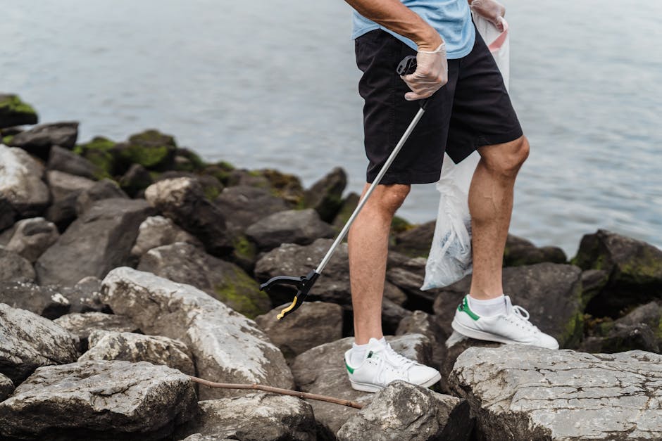 The image shows a person with a light blue t-shirt, black shorts, and white sneakers with green accents, standing on a rocky shoreline near a body of water, possibly a lake or river. The individual is holding a grabber tool in their right hand, which is extended towards the ground, and is wearing a disposable glove on the same hand. The rocks surrounding them are irregular in shape and size, with some having patches of moss or lichen, and vary in color from gray to dark brown. In the background, the water appears calm with gentle ripples, and the lighting suggests an overcast day, providing diffuse, soft illumination across the scene. The person's stance indicates they may be engaged in picking up litter or debris from the rocks, which ties into private waste removal or on-site cleanup activities that Rubbish Removal Chelsea offers as part of their rubbish services, highlighting a focus on outdoor rubbish clearance in natural environments, avoiding reliance on local authority collection. The image emphasizes environmental responsibility and private waste handling in outdoor, coastal settings. Keywords such as rubbish and waste management are naturally supported through the context of debris collection in a natural habitat.