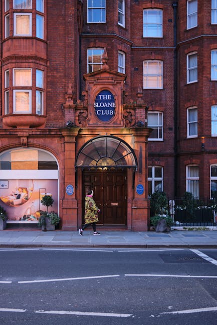 A woman wearing a floral dress and dark shoes is walking past the entrance of a red brick building with Victorian architectural features, including ornate stone detailing and multiple large sash windows. The entrance has a curved glass canopy above a wooden double door and is flanked by two blue signage plaques. Above the doorway, a blue sign reads 'The Sloane Club,' indicating a private members' club or venue. The ground-level window display to the left showcases a modern interior with a white chair, a side table, and decorative plants, providing a contrast to the historic exterior. The street in front has a clear, paved pavement with a curb separating it from the road, marked with white dashed lines. Small black wrought iron fences with potted plants border the building’s front, adding greenery to the scene. The lighting suggests late afternoon or early evening with a warm glow, underlining the building's red brick finish and textured stone accents, fitting within the context of independent waste management or rubbish removal services such as those offered by Rubbish Removal Chelsea, especially for flats or property management in the area.